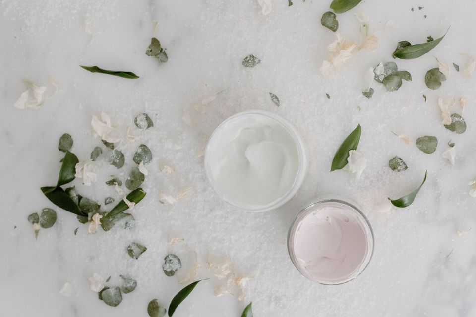 Dois potes abertos de creme sobre mármore branco, cercados de folhas e pétalas, em vista de cima