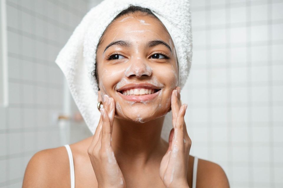 Mulher com toalha branca na cabeça lavando o rosto com espuma de sabonete em um banheiro de azulejos claros