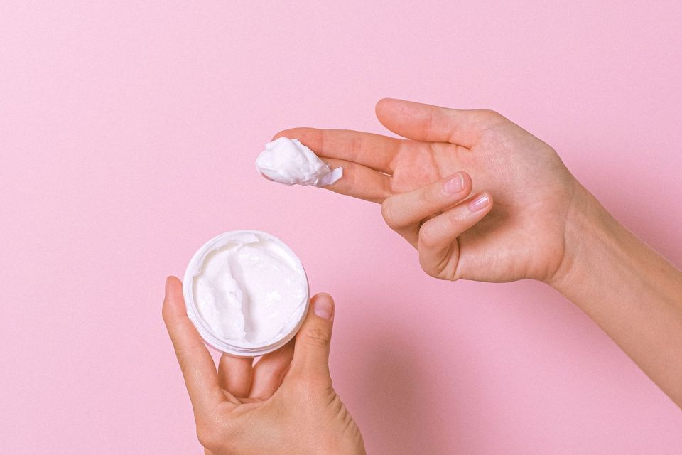 Mãos segurando um pote de creme branco e retirando um pouco do produto com os dedos, sobre fundo rosa