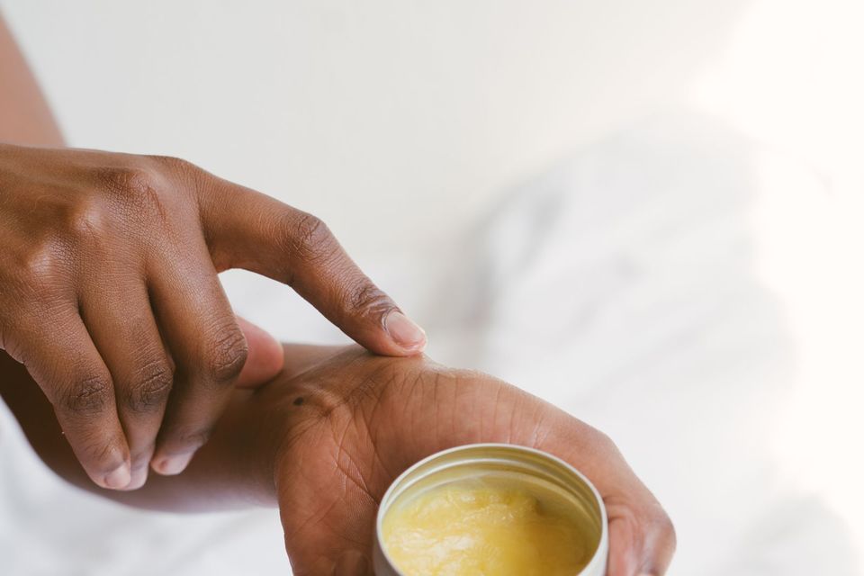 Mãos de uma pessoa segurando um pote aberto de creme e tocando o produto com o dedo