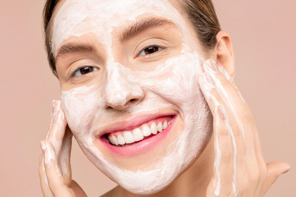 Mulher sorrindo com espuma de limpeza no rosto, com as mãos junto às bochechas, em fundo rosado