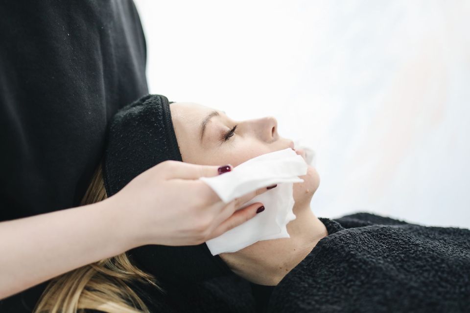 Profissional limpando delicadamente o rosto de uma mulher deitada, com faixa de cabelo preta, em ambiente claro