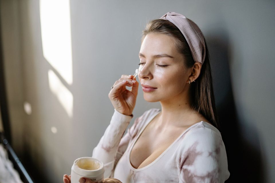 Mulher de faixa no cabelo aplicando sérum com conta-gotas no rosto, segurando um pote de creme, sob luz suave de janela