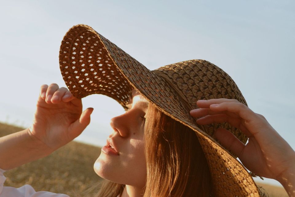 Mulher ruiva de camisa branca segurando a aba de um chapéu de palha largo e olhando para o sol, em um campo dourado no fim da tarde