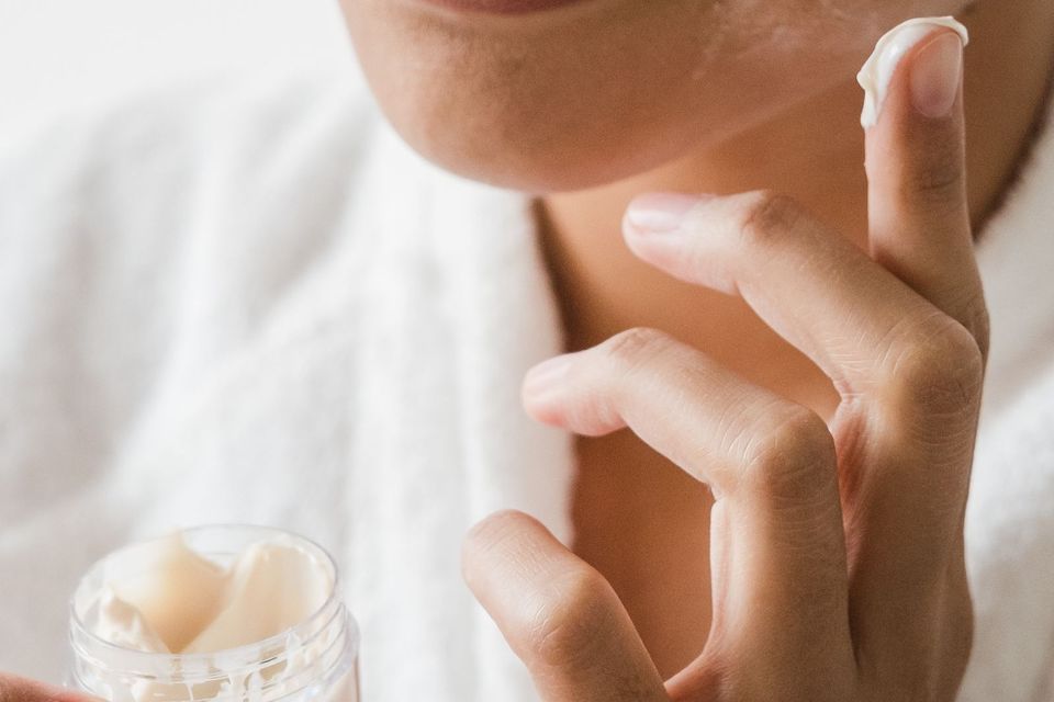 Mulher em roupão branco passando creme no rosto com o dedo, segurando um pote de vidro