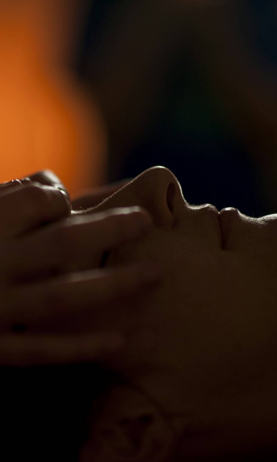 Mãos em massagem facial, em silhueta contra uma luz âmbar e quente